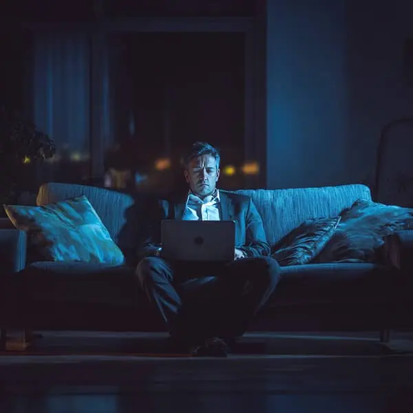 Homme d’affaires fatigué travaillant tard le soir sur son ordinateur portable dans un salon faiblement éclairé, illustrant la surcharge de travail et l’équilibre vie pro-vie perso.