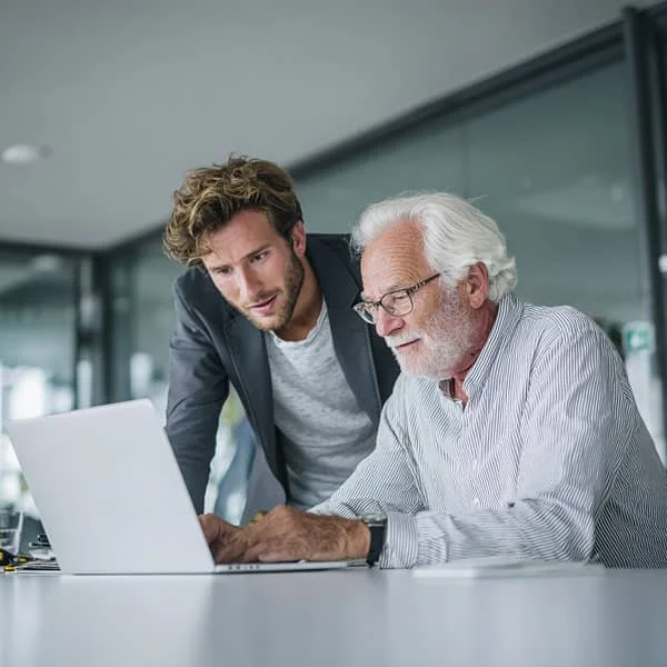 Jeune professionnel et collègue senior collaborant sur un ordinateur portable dans un bureau moderne, illustrant le mentorat intergénérationnel.