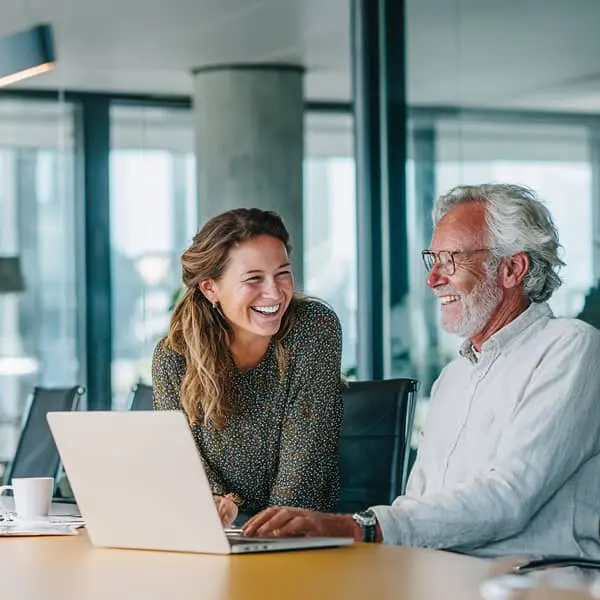 Jeune femme souriante expliquant quelque chose sur un ordinateur portable à un collègue plus âgé dans un bureau moderne, illustrant le concept de reverse mentoring et la collaboration intergénérationnelle.