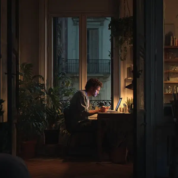 Trabajador en casa usando portátil y móvil de noche, ilustrando la falta de desconexión digital y la prolongación de la jornada laboral.