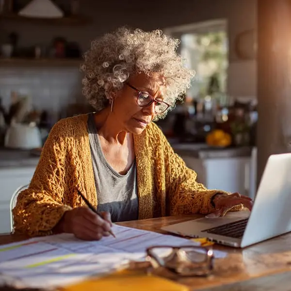 Femme senior assise à une table de cuisine, concentrée sur sa recherche d’emploi sur ordinateur portable, avec des documents et des notes étalés devant elle, éclairée par une lumière naturelle.
