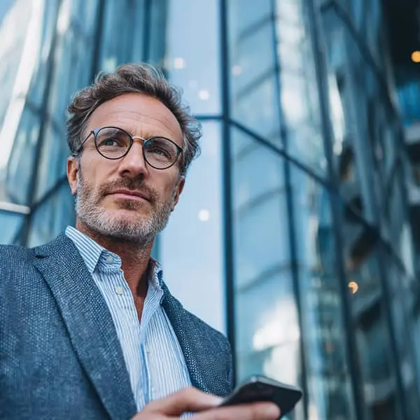 Cadre masculin en costume, debout en pleine journée devant une tour de bureaux vitrée, smartphone à la main, regard tourné vers l’horizon dans une ambiance business.