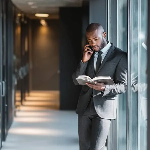 Recruteur en costume dans un couloir d’entreprise, appuyé contre une baie vitrée, parlant au téléphone tout en consultant un carnet de notes.