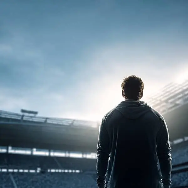 Athlète de haut niveau vu de dos, debout au bord d’un terrain dans un stade presque vide en pleine journée, symbole de transition et de reconversion après la carrière sportive.