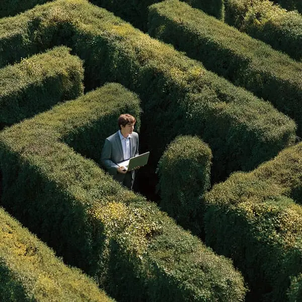 Professionnel en costume tenant un ordinateur portable au centre d’un labyrinthe végétal, symbolisant la difficulté de choisir un logiciel ATS