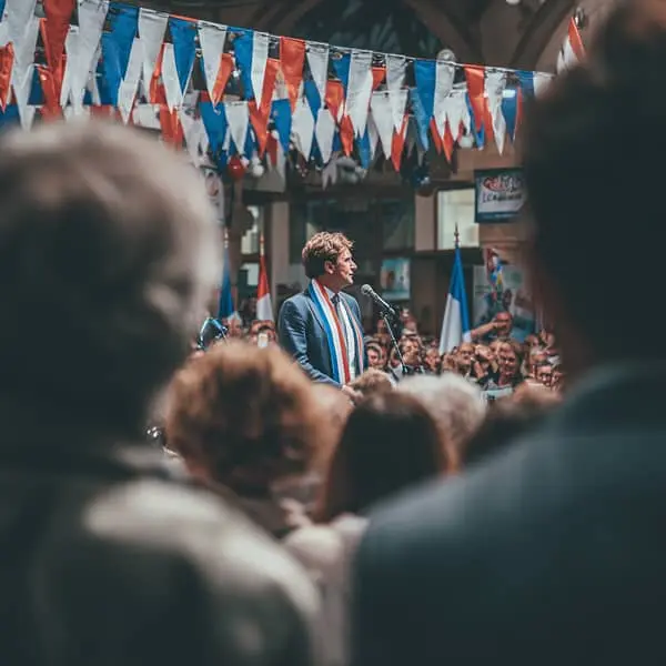 Image d'un maire s'exprimant lors d'un meeting public, avec des drapeaux tricolores en arrière-plan et une foule attentive. Le maire porte l'écharpe tricolore et se tient au micro, captivant l'attention des spectateurs.