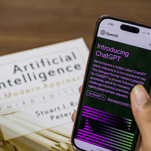 Photorealistic close-up of a hand holding a smartphone displaying an AI webpage, with an open artificial intelligence book blurred in the background, warm lighting, shallow depth of field, professional photography, ultra detailed.