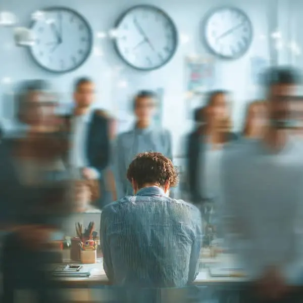 Salarié isolé et stressé au bureau, entouré de collègues flous et d’horloges symbolisant la pression et les risques psychosociaux au travail