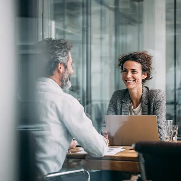 Entretien de recrutement dans une salle de réunion vitrée, une recruteuse souriante échange avec un candidat assis en face d’elle devant un ordinateur portable.