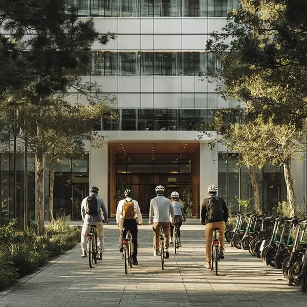 Groupe de salariés arrivant à vélo devant un immeuble de bureaux moderne, entouré d’arbres et de trottinettes en libre-service - illustration de la mobilité durable au travail.